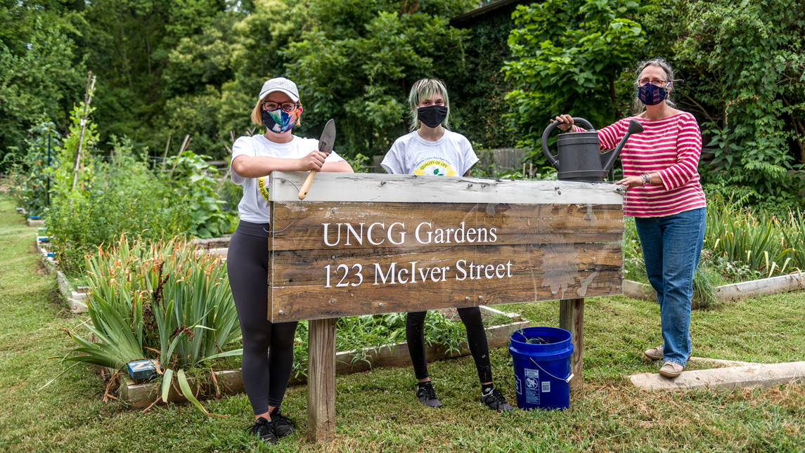 3 gardeners stand at the "UNCG Gardens" sign on McIver Street